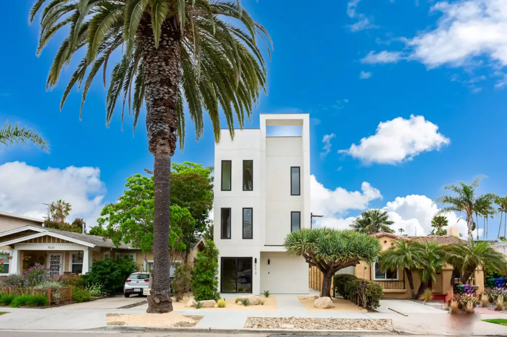 Modern white three-story house with tall windows, palm trees, and clear blue sky.